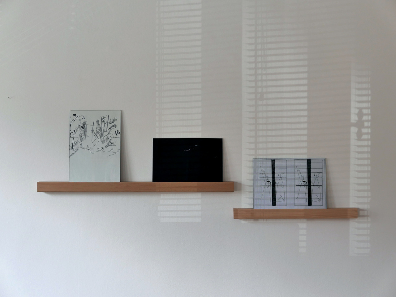 a photograph showing the view of the overall exhibit. the knight drawing, the dark photo and the character drawings are resting on tiny wood shelf against the wall. There is the shade of Venetian blind from the window behind letting some shape appears against the wall
